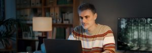 Mężczyzna ubrany w pasiasty sweter siedzi przy biurku i patrzy na laptopa przed sobą