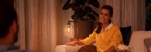 „Przytulna atmosfera salonu stworzona przez ciepłą lampę stołową LED, oświetlającą kobietę w żółtym swetrze cieszącą się wieczorną rozmową”.